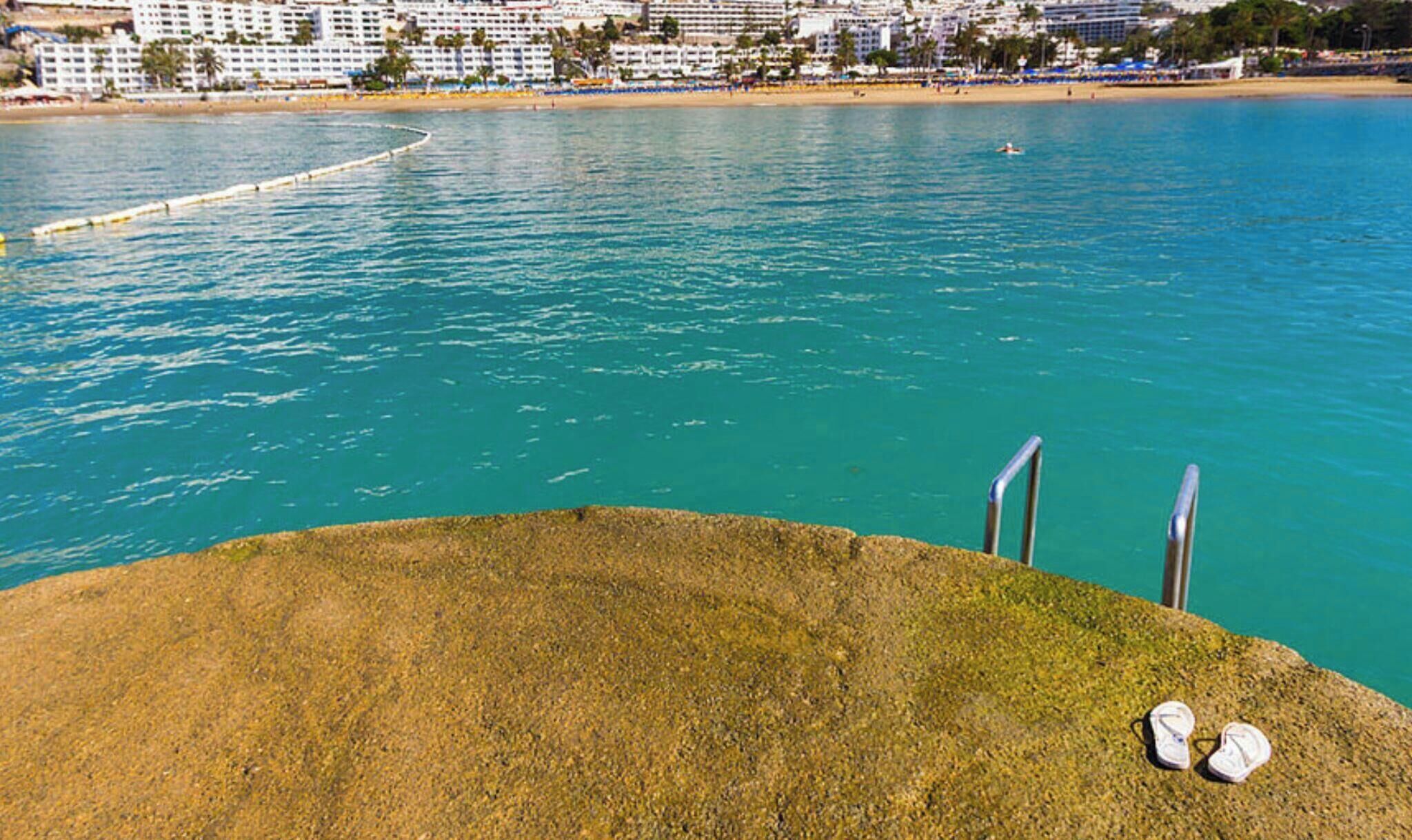 A coastal view with a clear blue sea, a sandy beach, and a cityscape in the background.