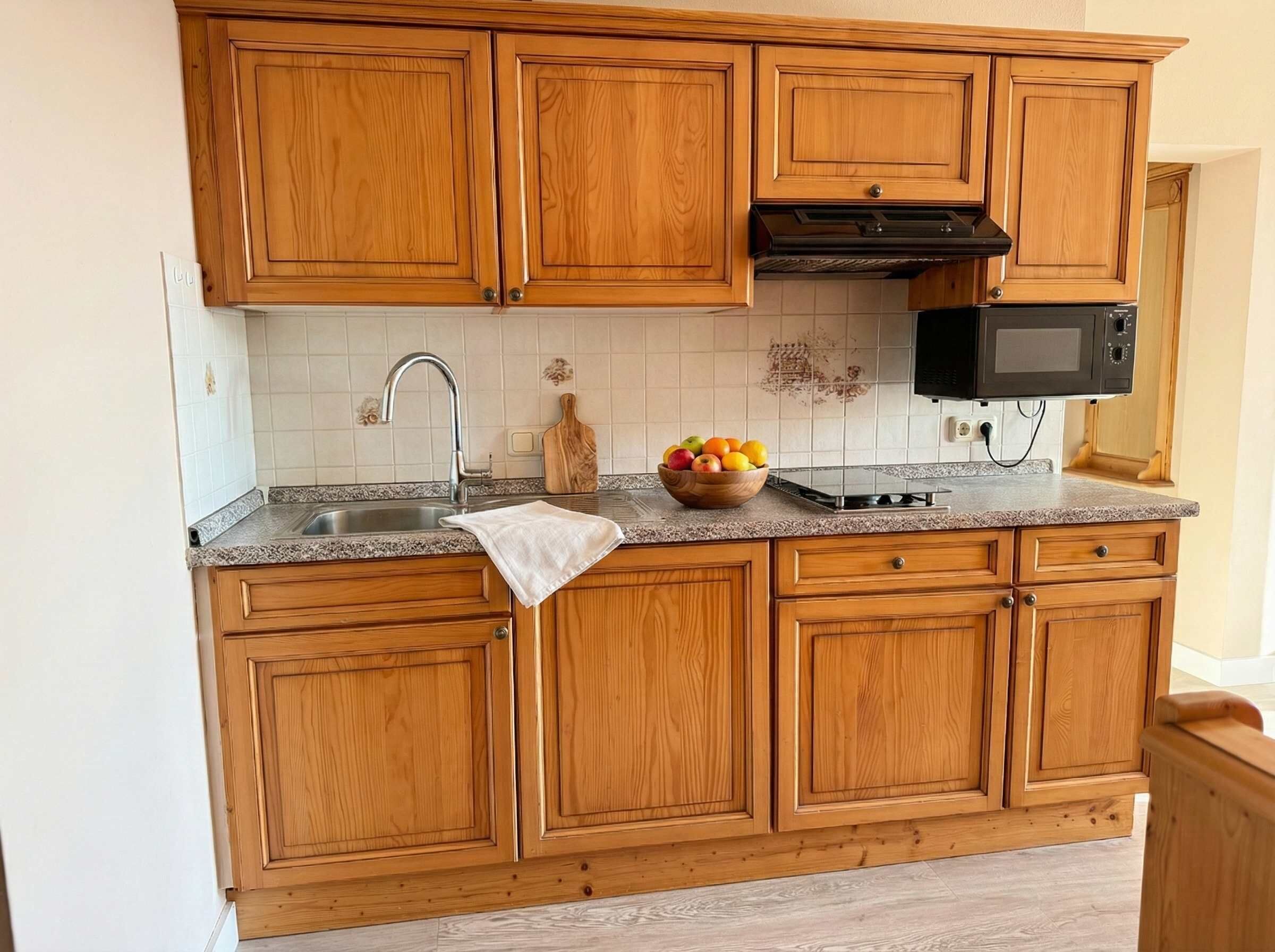 Keittiössä on puiset kaapit, ruostumattomasta teräksestä valmistettu pesuallas, mikroaaltouuni ja hedelmäkulho työtasolla.