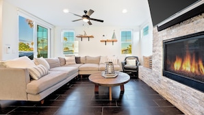A modern living room with a sectional sofa, a round coffee table, a fireplace, and a large TV screen. - Grand 5BD Tri-Level Home with Bay Views Garage Parking AC & Large Patio! (San Diego)