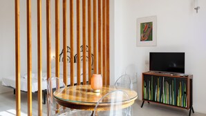 Un soggiorno moderno con un tavolo rotondo in vetro, sedie trasparenti, un pannello divisorio in legno, un mobile TV e un letto sullo sfondo.