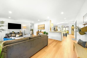 A modern living room with a sofa, a TV, and a dining area with a kitchen in the background. - Amazing Reviews - Large Back Yard, Firepit W/AC, Stunning! (San Diego)