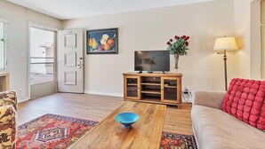 Una sala de estar con una mesa de centro de madera, un televisor sobre un mueble de madera, un cojín rojo en un sofá y una alfombra colorida.