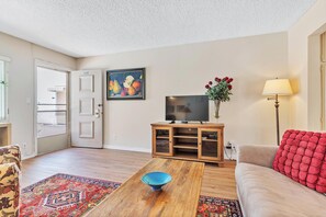A living room with a wooden coffee table, a television on a wooden stand, a red cushion on a sofa, and a colorful rug. - Perfect Location W/Pool and Parking, Walk 2 Beach! (San Diego)