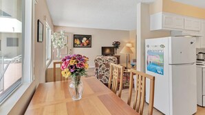 Un comedor con una mesa de madera, sillas, un florero con flores, un televisor y un refrigerador.