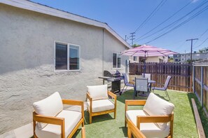 Un jardin avec un barbecue, deux chaises en bois, une petite table et un parasol violet.