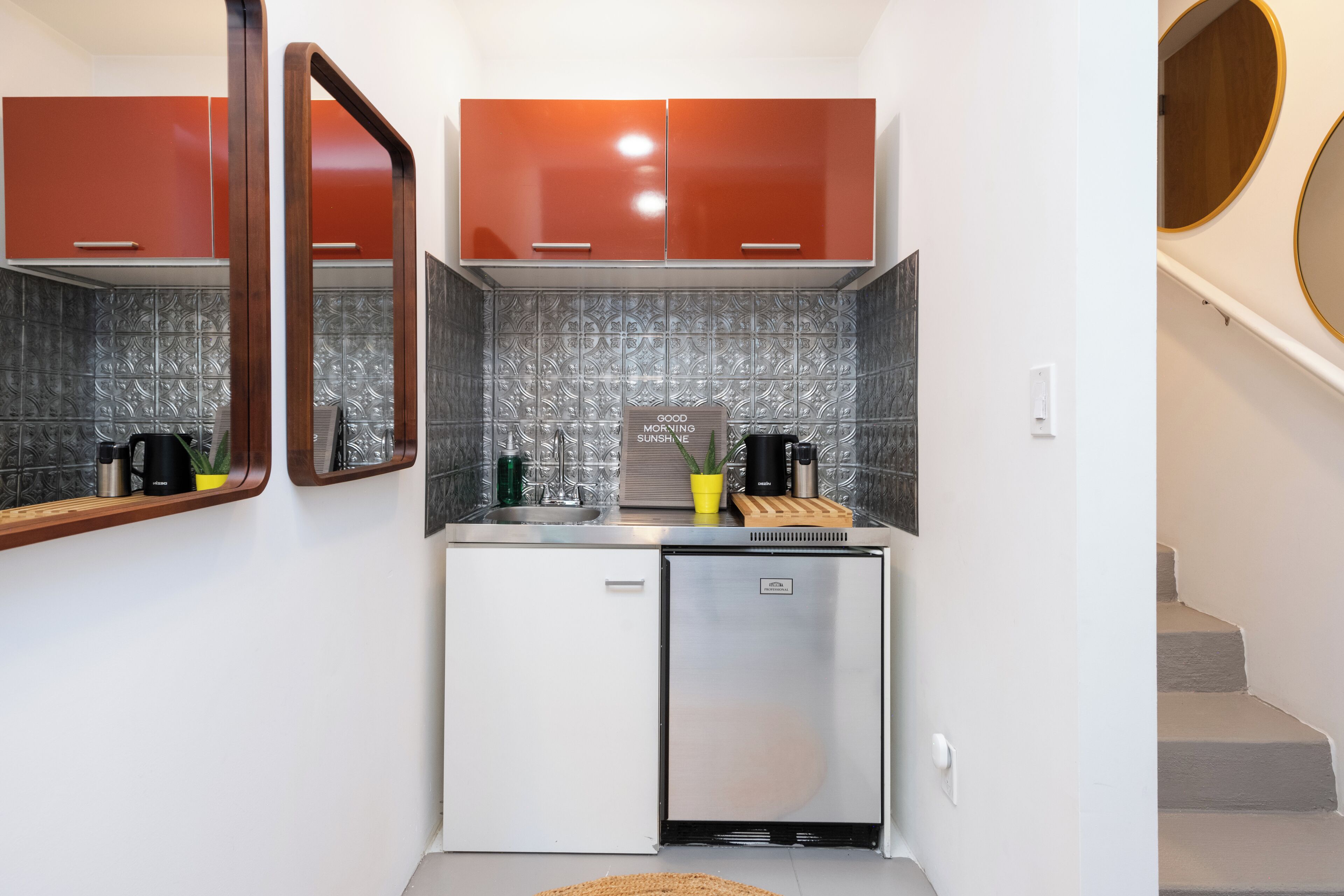 Uma cozinha compacta com geladeira, pia e bancada com diversos eletrodomésticos e uma planta em vaso.