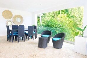 A modern outdoor dining area with wicker furniture, a glass balustrade, and a view of lush greenery. - Brisa Azul | Poolfront | Walk to Beach | Wi-Fi (Las Terrenas)
