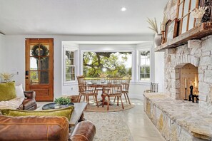 A cozy living room with a stone fireplace, a dining area with a round table and chairs, and a large window offering a view of greenery. - Blanco Hill Country Hideaway (Blanco)
