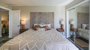 Un dormitorio con una cama grande, cabecera decorada, dos mesitas de noche con lámparas y un espejo en la pared.