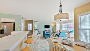 A modern kitchen with a marble countertop, a dining area with a wooden table and white chairs, a blue sofa, and a TV mounted on the wall. - Perfect For Families, Steps To Beach, Close to Everything! (San Diego)