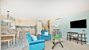 A living room with a blue sofa, a coffee table, a TV, and a kitchen area with white cabinets. - Perfect For Families, Steps To Beach, Close to Everything! (San Diego)