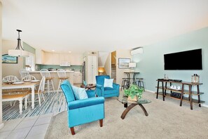 A living room with a blue sofa, a coffee table, a TV, and a kitchen area with white cabinets. - Perfect For Families, Steps To Beach, Close to Everything! (San Diego)