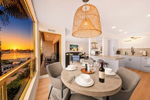 A dining area with a view of the sunset, a round table set with plates, wine, and cutlery, and a kitchen in the background. - Pet Friendly 2 bedroom in Point Loma Overlooking Kellogg Beach - Stay for 30 days! (San Diego)