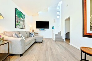 A living room with a sofa, a flat-screen TV, a staircase, and a painting on the wall. - Great Reviews - Steps 2 Beach W/Garage & Parking! (San Diego)