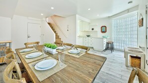 Un comedor con una mesa de madera preparada para cuatro personas, una cocina al fondo y una escalera que sube a un nivel superior.