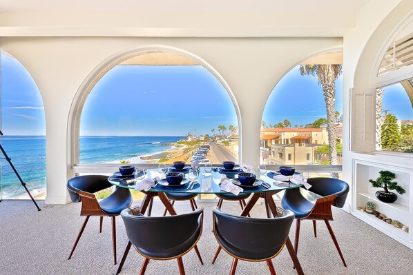 Ein Essbereich mit Tisch für vier Personen, mit Blick auf Strand und Meer durch bogenförmige Fenster.