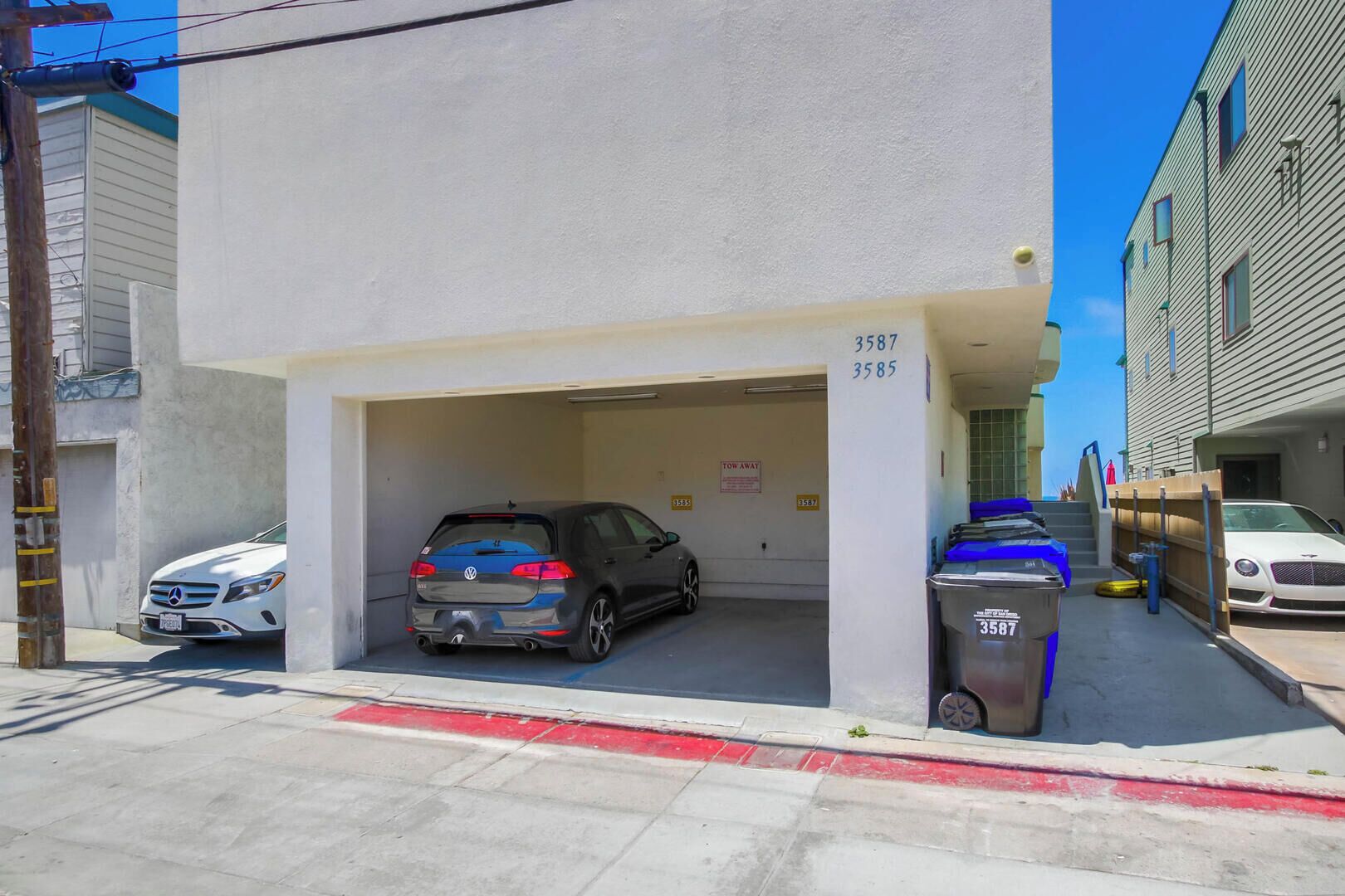 A parking garage with two cars parked inside, a white building with a garage door, and a trash can labeled 3587.