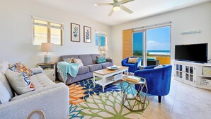 Una sala de estar con un sofá gris, sillones azules, una mesa de centro y un televisor. Cuenta con un ventilador de techo, un ventanal con vista al océano y una decoración temática de playa.