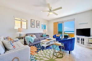 A living room with a grey sofa, blue armchairs, a coffee table, and a TV. There is a ceiling fan, a large window with a view of the ocean, and a beach-themed decor. - Beachfront, Endless Sunset Views, Amazing Location W/Parking! (San Diego)