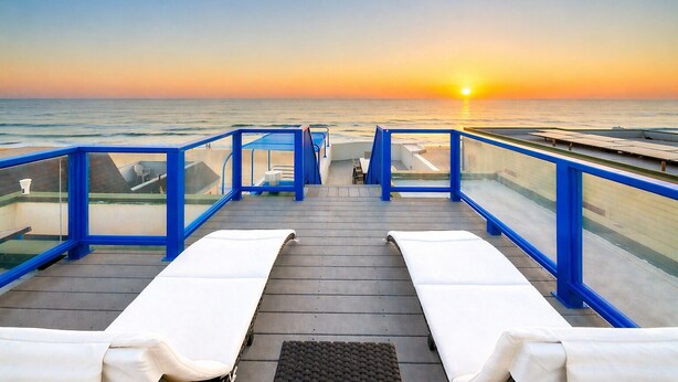 Une terrasse dotée de deux chaises longues blanches, d’une balustrade en verre et offrant une vue sur le coucher de soleil sur l’océan.