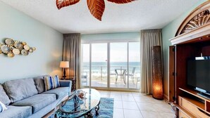 Ein Wohnzimmer mit einer Couch, einem Couchtisch und einem Fernseher, ein großes Fenster mit Blick auf den Strand und eine Decke mit Blattverzierungen.