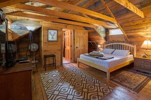 A cozy attic bedroom with a wooden ceiling, a bed with patterned bedding, a desk with a computer, and a standing fan.