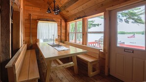 A wooden cabin interior with a dining table, benches, and large windows offering a view of a lake and dock. - Waterfront Victoria Cottage,Kayaks,paddle board and canoe Included, Dogs Welcome (Clifton)