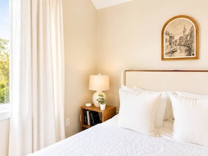 A hotel room with a bed, a nightstand with a lamp, a framed picture on the wall, and a window with curtains.
