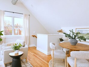 A modern living room with a sloped ceiling, a white sofa, a round wooden table, and a view of the outside.
