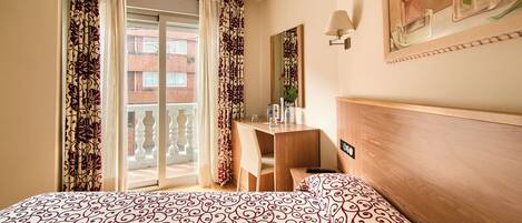 A hotel room with a bed, a desk, a chair, and a balcony with curtains.