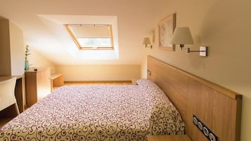 Un dormitorio con cama, escritorio, silla, mesita de noche, lámpara, un cuadro en la pared y una ventana.