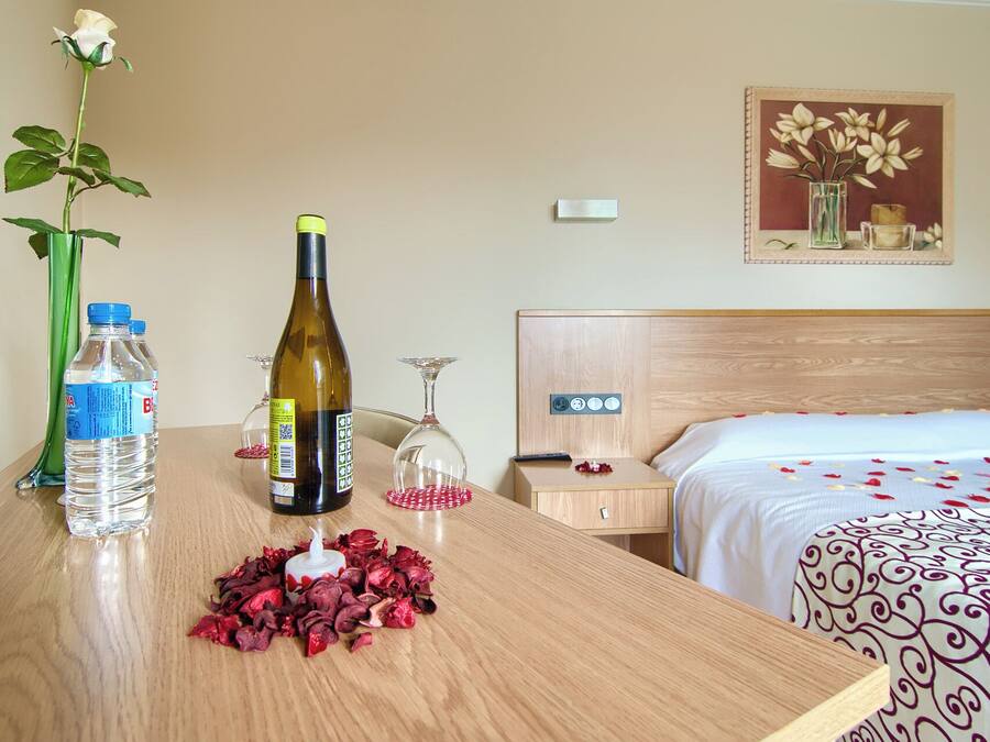 A hotel room with a wooden bed, a bedside table with a bottle of water, a bottle of wine, a candle, and a vase with a single white rose.