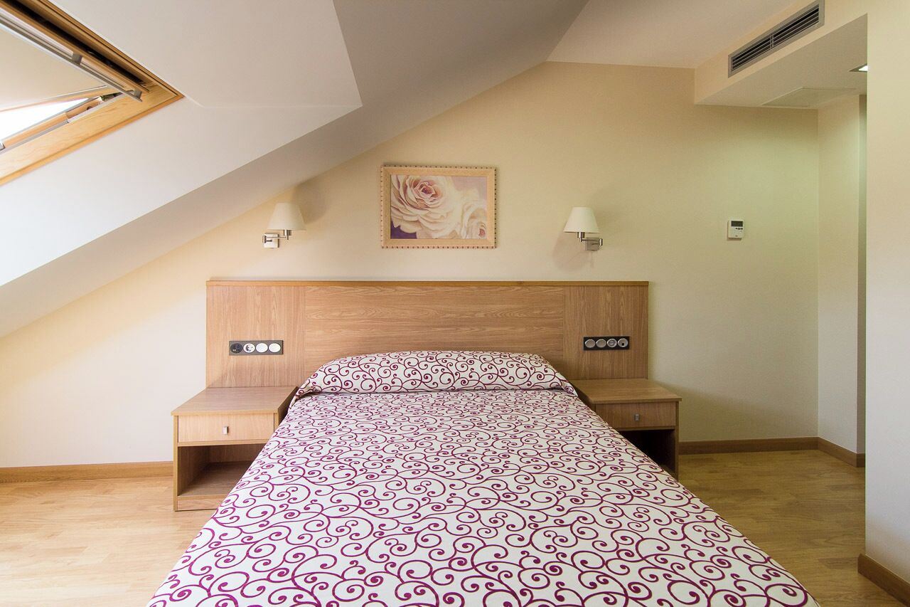 A bedroom with a sloped ceiling, a bed with a patterned bedspread, two bedside tables, a wall-mounted light, and a framed picture above the bed.