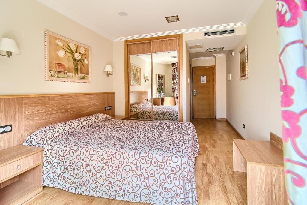 A hotel room with a wooden headboard, a patterned bedspread, a wooden nightstand, a mirror, and a door leading to a bathroom.
