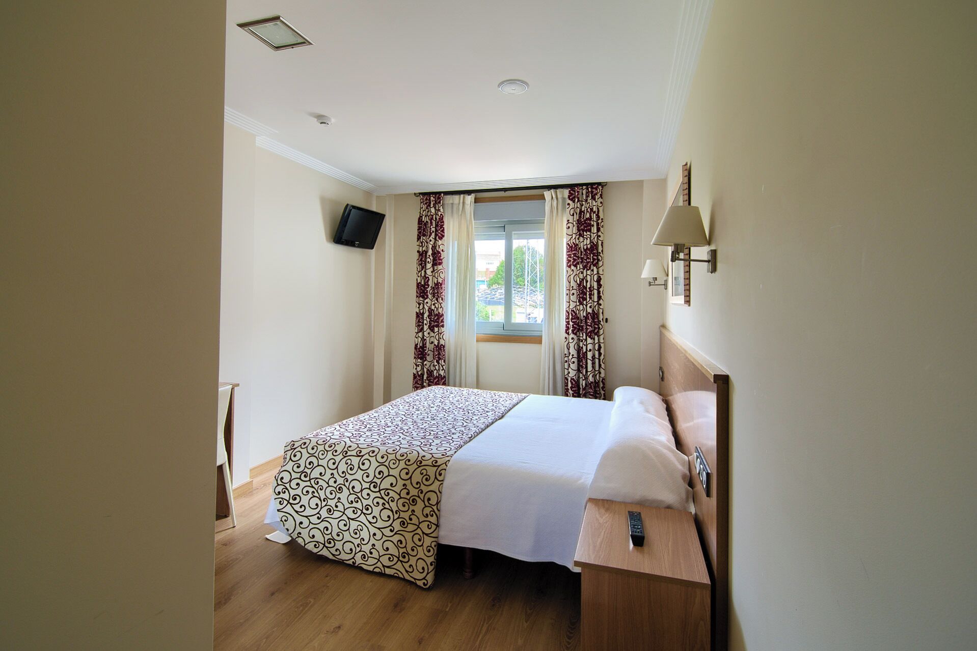 Una habitación de hotel con una cama, televisor en la pared, ventana con cortinas y una lámpara de noche.
