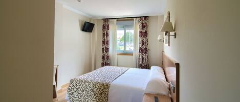 Una habitación de hotel con una cama, televisor en la pared, ventana con cortinas y una lámpara de noche.