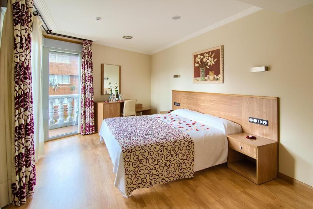 Habitación de hotel con una cama grande, cabecera de madera, mesitas de noche, un espejo y vistas al balcón exterior.
