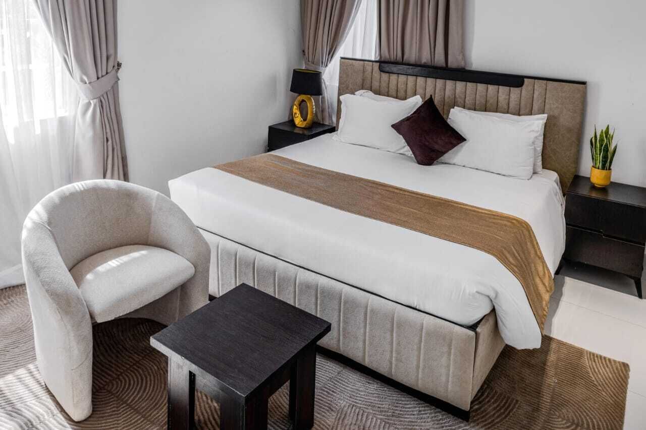 A hotel room with a large bed, a chair, a small table, and a nightstand with a lamp.
