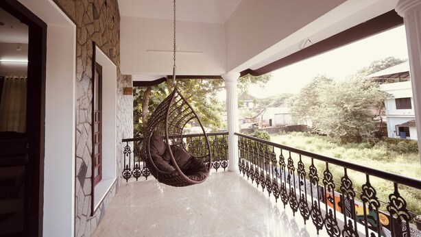 A balcony with a hanging chair, a stone wall, and a view of greenery and buildings. - 3-bedroom house in peaceful Alappuzha with AC comfort (Alappuzha)