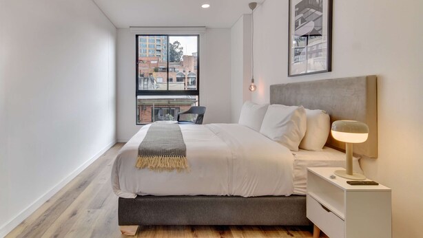 Quarto moderno com cama grande, mesa de cabeceira com luminária, vista de edifícios pela janela e um quadro na parede.
