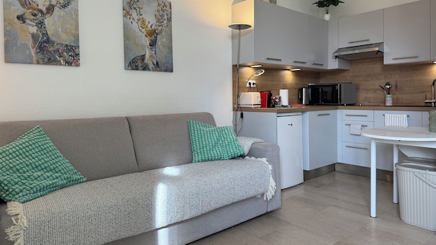 Ett modernt kök med vita skåp, ett matbord och en soffa med gröna och vita kuddar. Det hänger två tavlor på väggen och en liten växt står på bänkskivan.