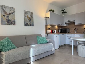 Une cuisine moderne avec des meubles blancs, une table à manger et un canapé agrémenté de coussins verts et blancs. Deux tableaux ornent le mur et une petite plante est posée sur le comptoir.