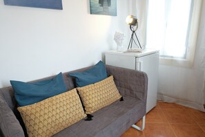Een grijze bank met twee gekleurde kussens, een klein wit nachtkastje met een lamp, en een raam met dunne gordijnen.