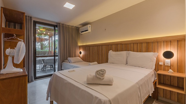A hotel room with a bed, bedside table, fan, and a view of the outdoors. - Wai Wai Cumbuco Residence #c101 by Carpediem (Caucaia)