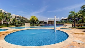 A swimming pool area with a central circular pool, surrounded by a stone deck, palm trees, and a building in the background. - Wai Wai Cumbuco Residence #c101 by Carpediem (Caucaia)