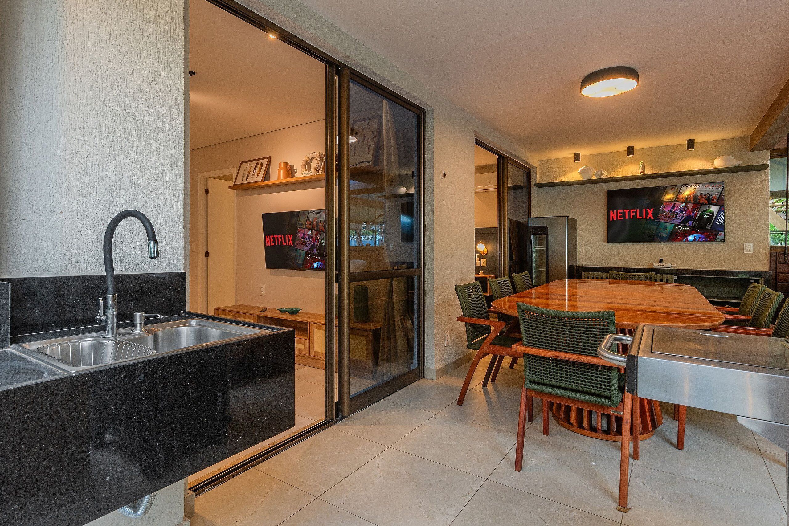 Una cocina moderna con un lavaplatos empotrado, una zona de comedor con una mesa y sillas de madera, y un televisor en la pared mostrando el logotipo de Netflix.
