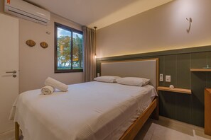A neatly made bed with a rolled towel, a bedside table, a wall-mounted shelf, and a window with curtains. - Wai Wai Cumbuco Residence #c101 by Carpediem (Caucaia)