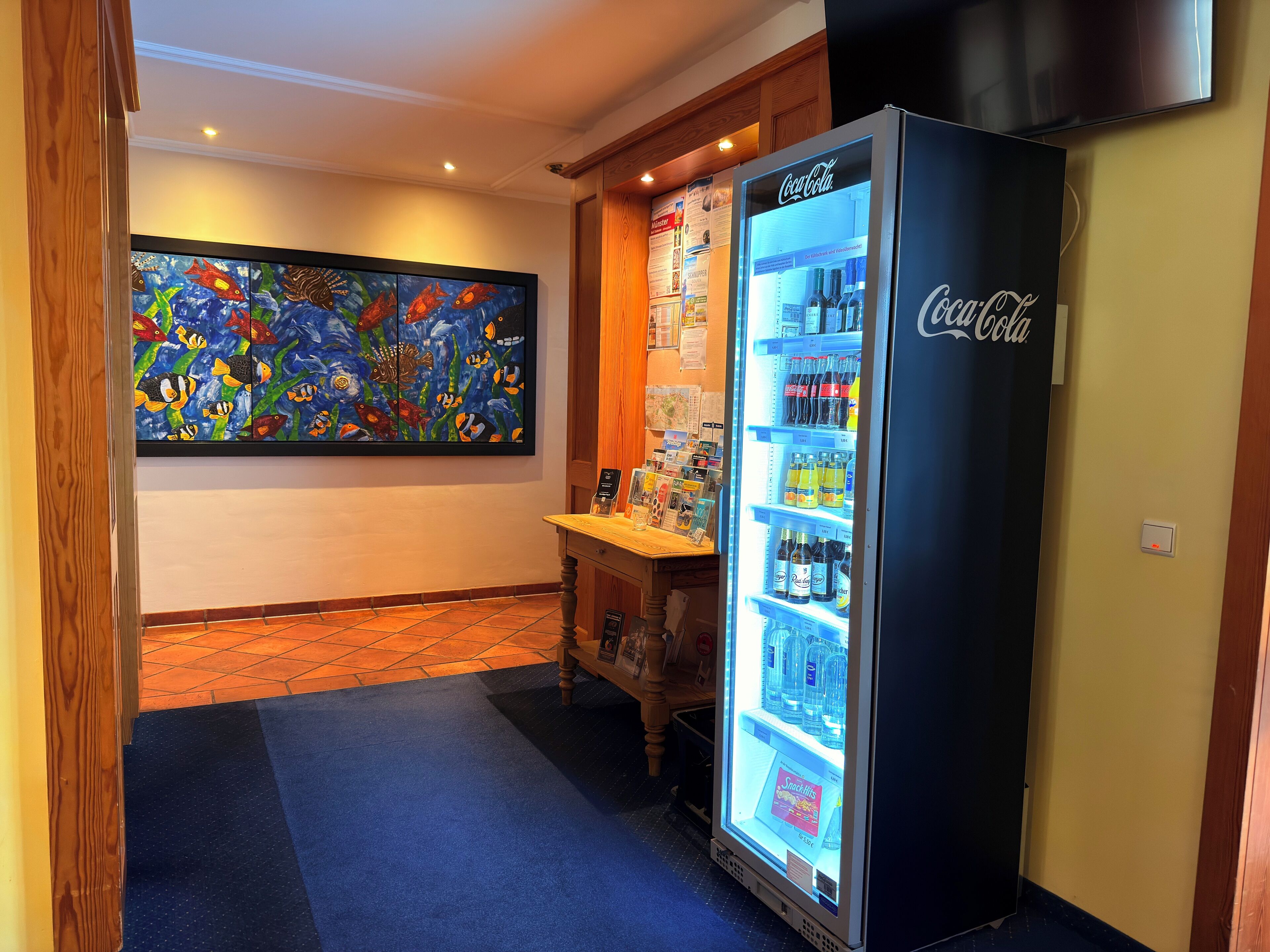 Un hall de hotel con un refrigerador de Coca-Cola, un cuadro de un pez colorido y una barra de madera con diversos objetos.