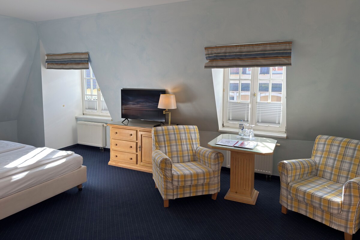 A bedroom with a bed, two plaid armchairs, a wooden dresser, a television, and a table with a lamp.