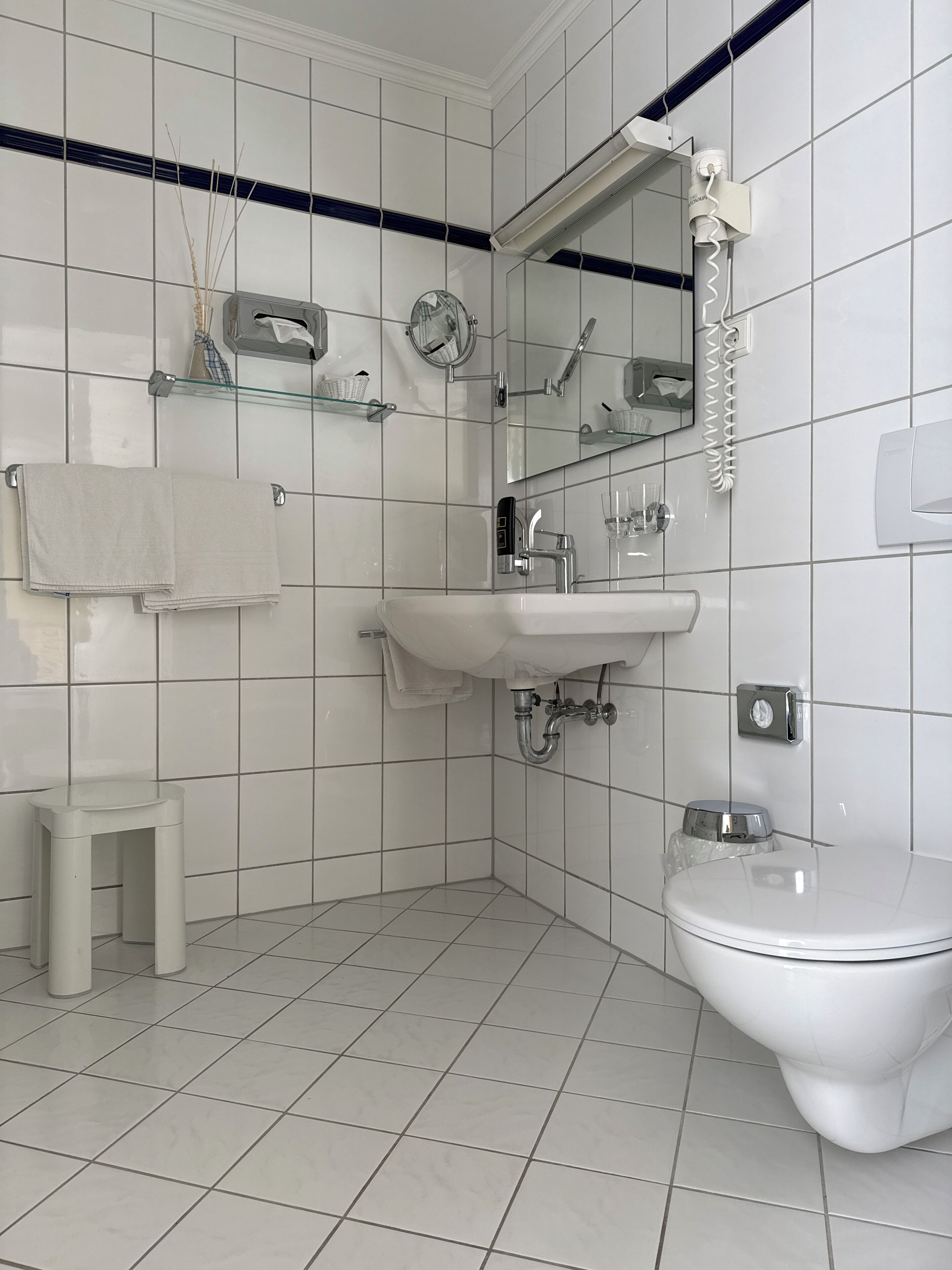 Une salle de bain avec des carreaux blancs, un lavabo, des toilettes, un miroir et une étagère en verre sur laquelle est posée une serviette.